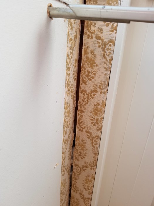 Bred spricka i vägg bakom delvis riven, mönstrad tapet med synliga spackelrester nedanför.