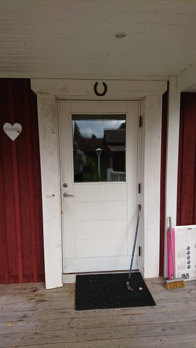 Nyinstallerad vit ytterdörr med fönster i en röd vägg, verktyg och borste på en träveranda.