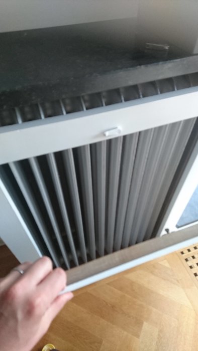 Hand som öppnar ett vitt elementskydd som avslöjar en traditionell radiator framför en fiskbensparkett.