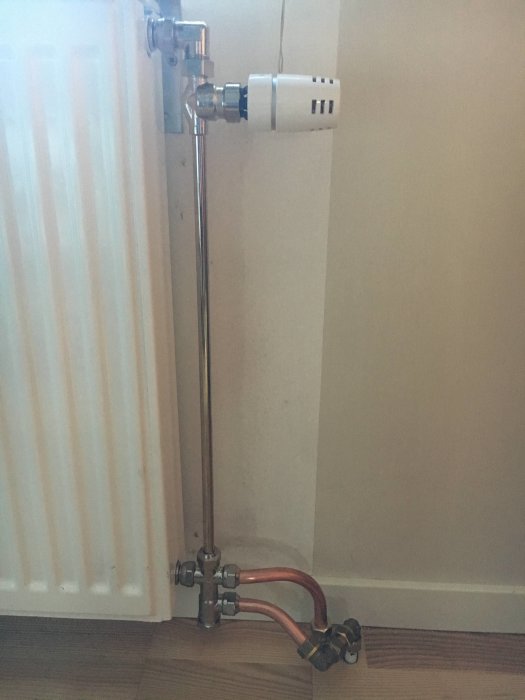 Radiatoranslutning i ett hörn med termostatventil och kopparledningar mot parkettgolv.