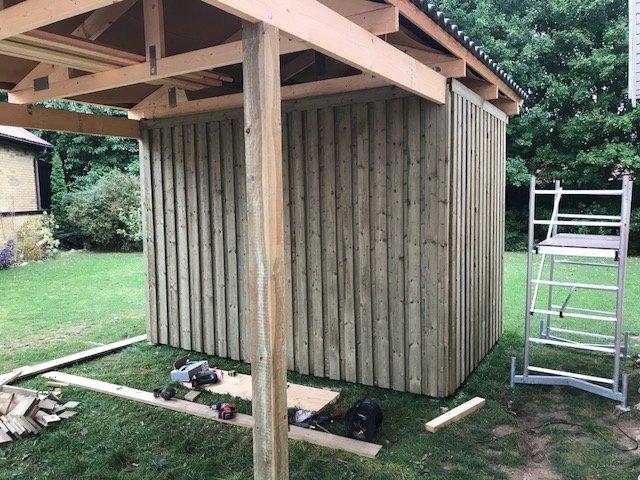 En nästan färdigbyggd träkonstruktion med vertikal panel under tak med verktyg och material på marken.