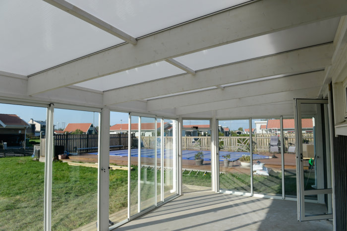Nybyggd veranda med tak och glaspartier som visar utsikten mot en trädgård med pool.