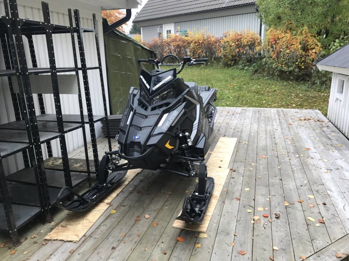 En snöskoter parkerad på en trätrall nära några metallhyllor och höstlöv på marken.