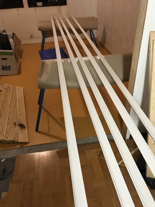 Vitmålade lister som ligger på ett gult golv under renovering med byggmaterial runtomkring.