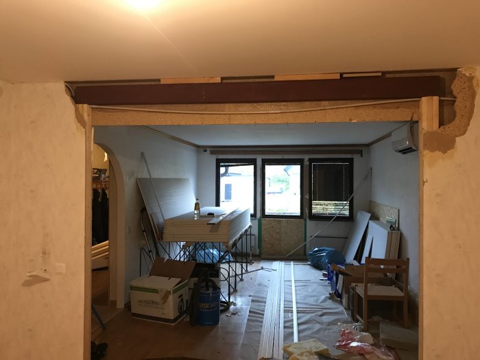 Renoveringsarbete pågår i hus med synlig bärbalk, byggmaterial och isolering i rum med bågformad öppning.