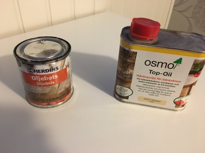 Två burkar med träbehandlingprodukter, Herdins Oljebets och Osmo Top-Oil, på en vit yta.