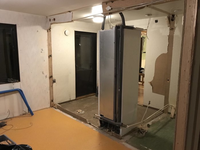 Renoveringsarbete inomhus med avlägsnade väggsektioner, synlig varmvattenberedare och gammal plastmatta.
