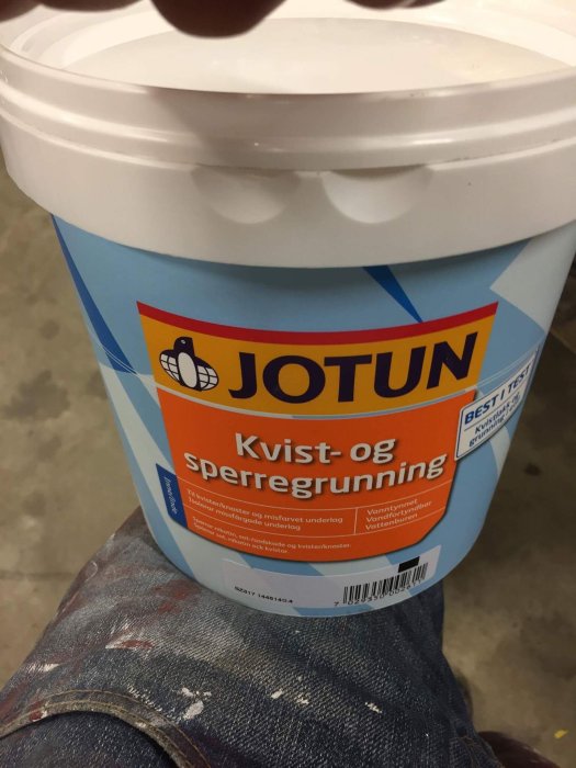 Burk med Jotun kvist- och spärrgrundfärg hållen i en hand med målarfärgfläckar på byxorna.