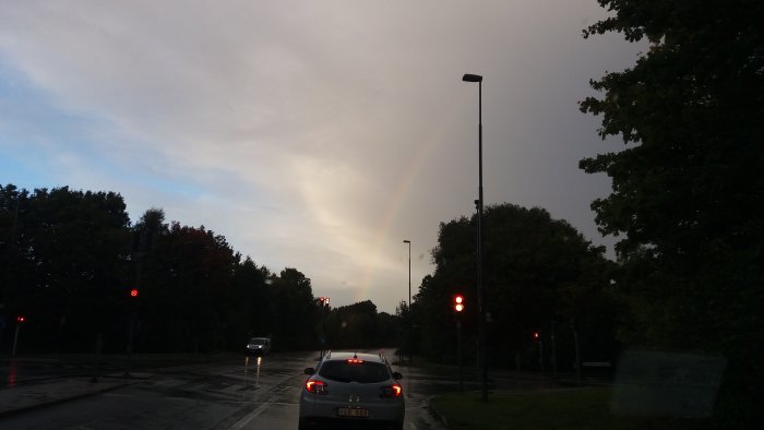 Regnvåt gata med bil och trafikljus, delvis solig himmel med regnbåge i bakgrunden.