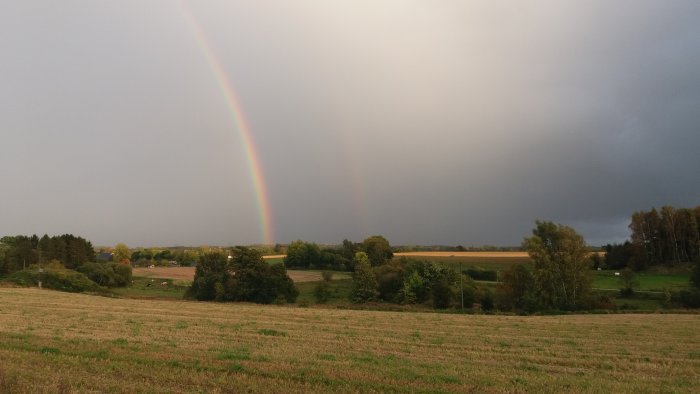 Dubbel regnbåge över ett höstfärgat landskap med molntunga himmel.