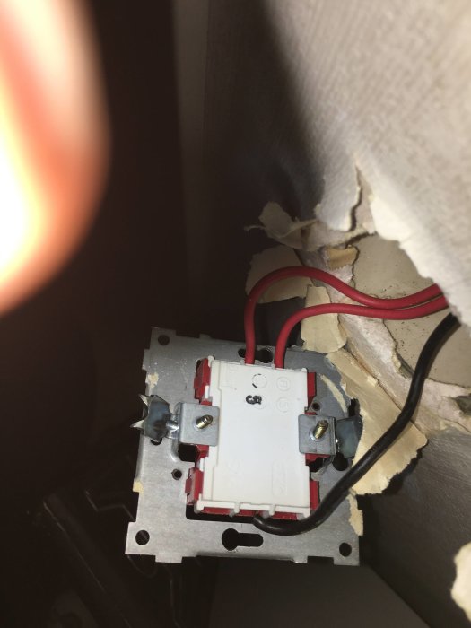 Elektrisk väggströmbrytare med två röda kablar anslutna ovanifrån och en svart, vid en trasig vägg.