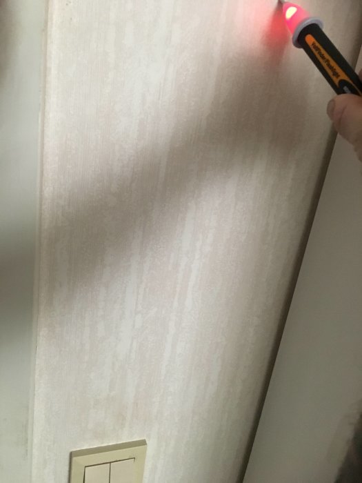 Hand som håller en spänningsprovare mot vägg ovanför en ljusbrytare, spänningsprovaren lyser.