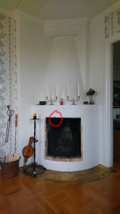 Vit hörnspis med öppen spis och mantel med ljusstakar och blomvas, omgiven av tapet och trägolv.