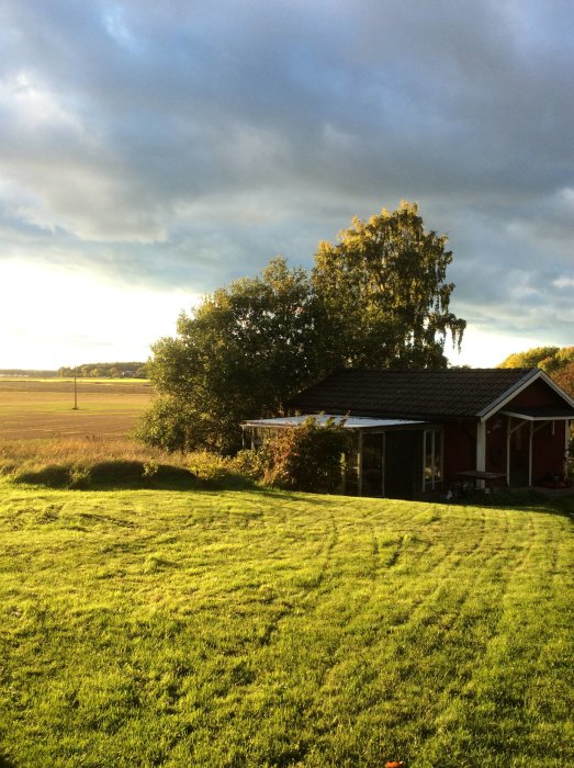 Eftermiddagssol över en röd villa med veranda i Ärsta, Sörmland, med öppet fält i bakgrunden.