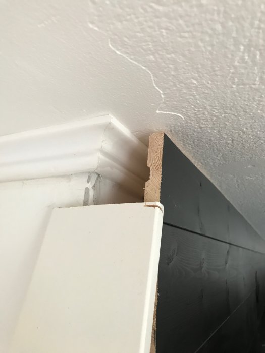 En hörndetalj där taklisten i frigolit möter en grundare foderlist ovanför en mörk panelvägg.