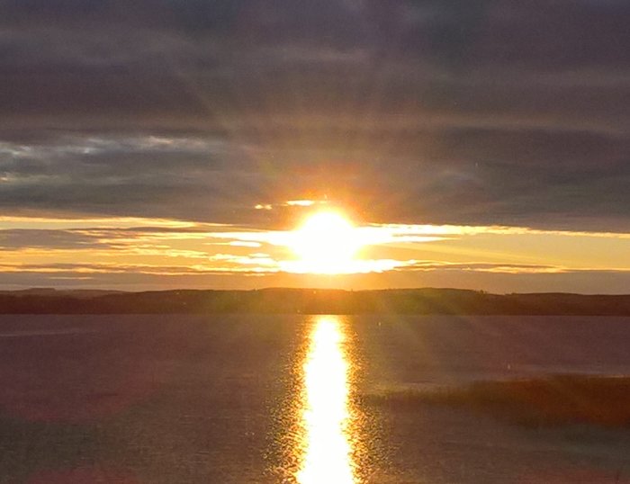 Soluppgång över ett stilla vatten med reflektion av solen på ytan och moln på himlen.