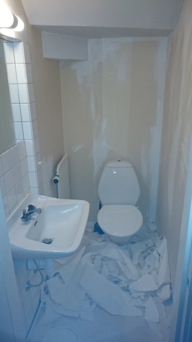 Ett badrum halvvägs genom renovering med nedriven tapet och spacklade väggar, inklusive handfat och toalett.