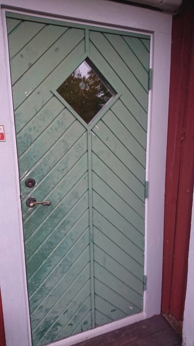 Grön ytterdörr med rombformad glasruta som hänger ner sig på framkanten, behov av justering.