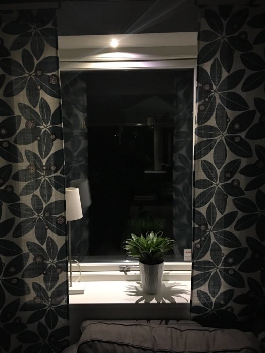 Inredningsbild med blommande gardiner, fönsterbänk med planta och lampa, reflekterande nattfönster.