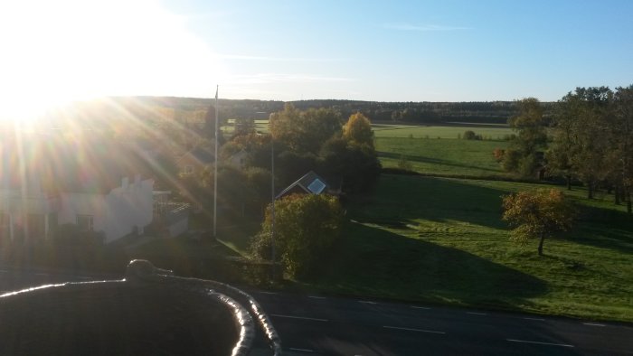 Vy från en taktopp över ett landskap med gröna fält, träd och hus i morgonsol.