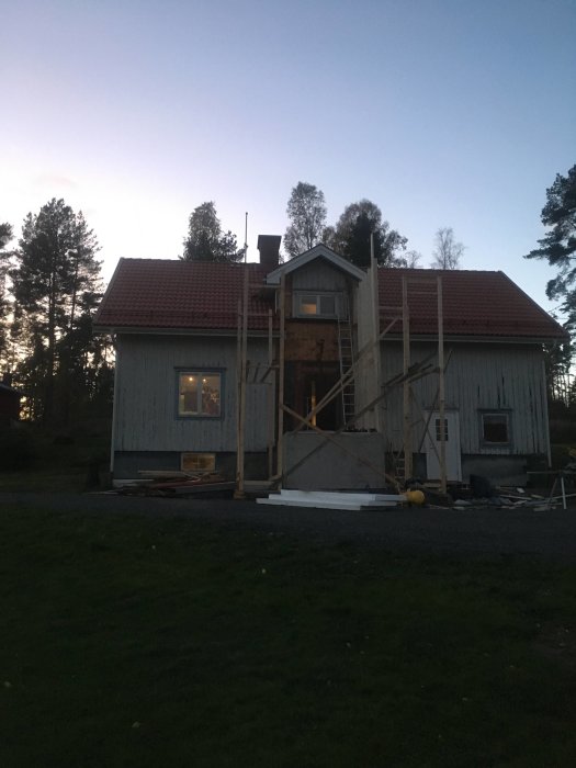 Hus under renovering med improviserad plankställning och byggmaterial på marken i skymningen.