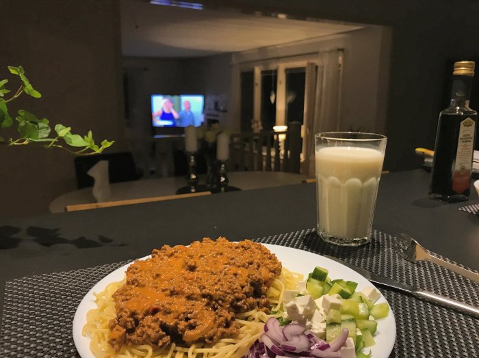 Måltid med spaghetti och köttfärssås på tallrik framför dimmigt kök och TV i bakgrunden.