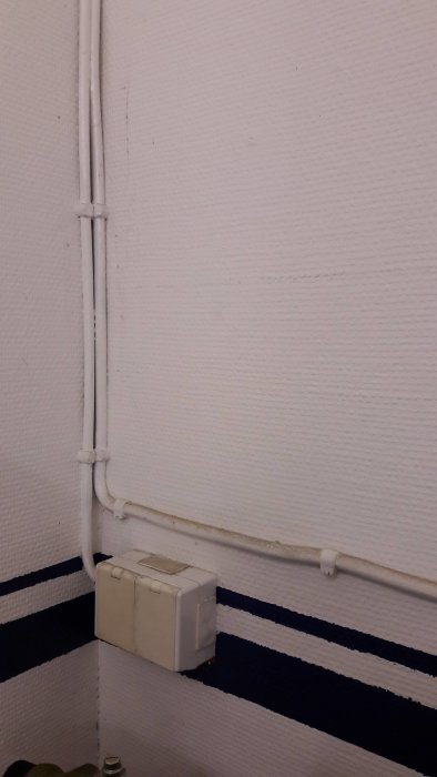 Äldre elektrisk central med automatsäkringar och trefasbrytare på texturerad vit vägg med svart och blå rand.
