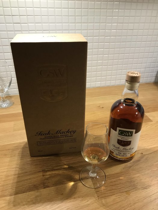 Flaska och förpackning av Isak Mackey Single Malt Whisky från Gammelstilla, med glas på träbord.