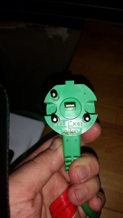 Grön plastkomponent med texten "SAMHALL K61P" hållen i en hand.