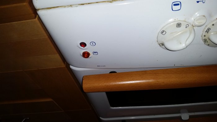 Närbild av en använd vit spisfront med reglage och en lampa som inte sitter fast korrekt.