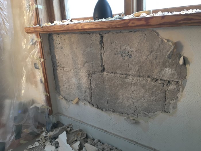 Avskalad vägg under fönster med synlig puts som delvis släppt från ytterväggen och bitar av rivna tapeter.