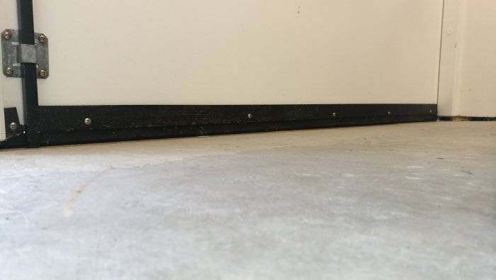 En fastklämd svart list längs med en vit vägg, säkrad med skruvar och ett metallbeslag, mot ett betonggolv.