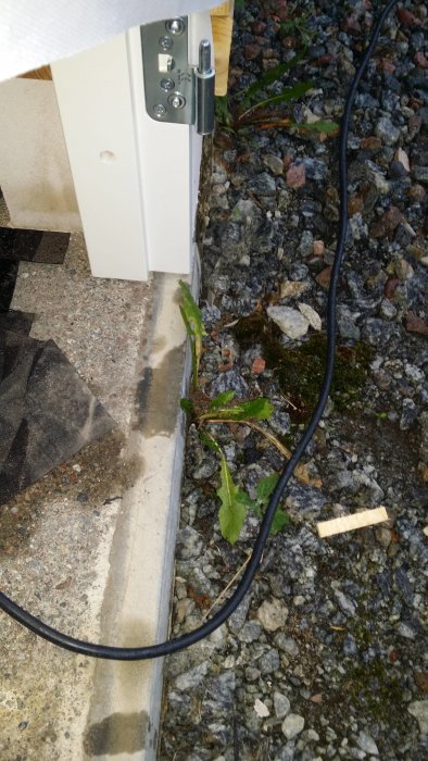 Detalj av en husgrund med vitt plåtarbete, betongkant och gruskant, samt en svart sladd som löper längs marken.