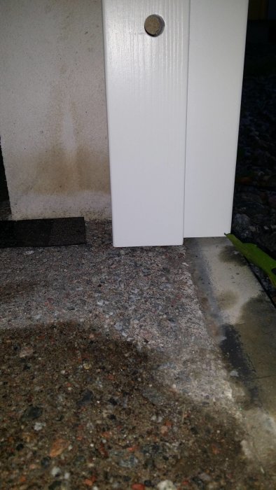 Vit dörrstolpe mot en betongvägg med fuktskador och grusig mark.