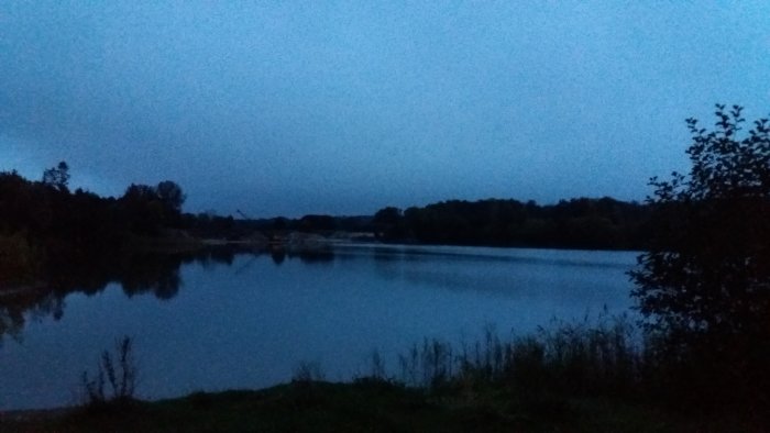 Kvällsutsikt över en stilla sjö med speglande vattenyta och mörka siluetter av träd.