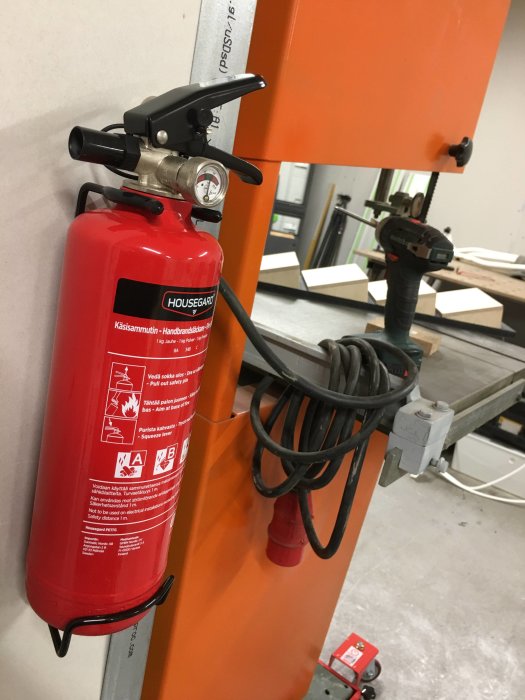 Montering av en 1kg röd brandsläckare på en orange pelare i en verkstad, med verktyg i bakgrunden.