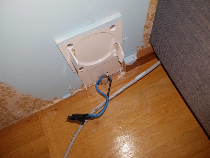 En öppen elektrisk vägguttag med oskyddade elkablar och borttaget lock vid en väggkant.
