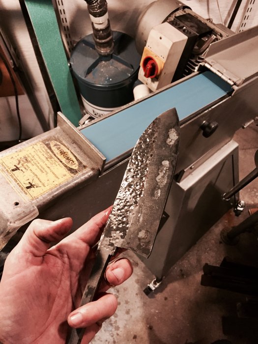 Hand håller ett nyslipat knivblad med grov yta efter slipning med 40 grit, framför slipmaskiner.