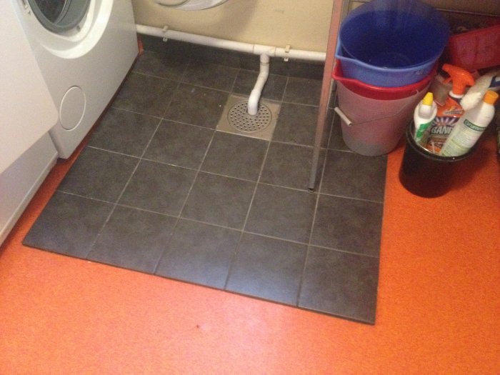 Kakelplattor limmade över orange matta runt brunnen i ett tvättstuga, med hinkar och rengöringsmedel synliga.