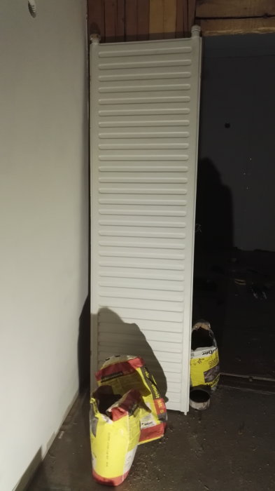 En vit radiator monterad på en vägg ovanför två säckar byggmaterial.
