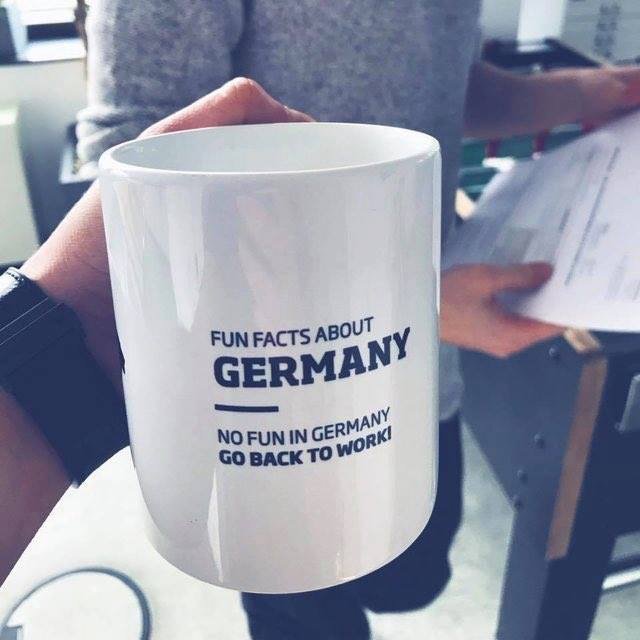 En person håller en vit kaffemugg med texten "Fun Facts About Germany - No Fun in Germany Go Back to Work".