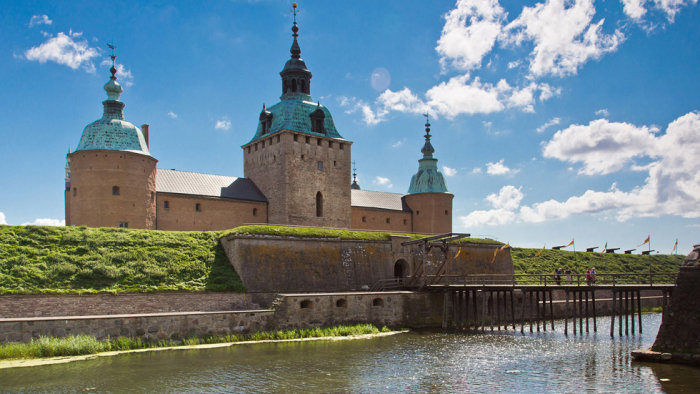 Ett historiskt slott med torn och kreneleringar omgivet av vatten och en träbro under blå himmel.