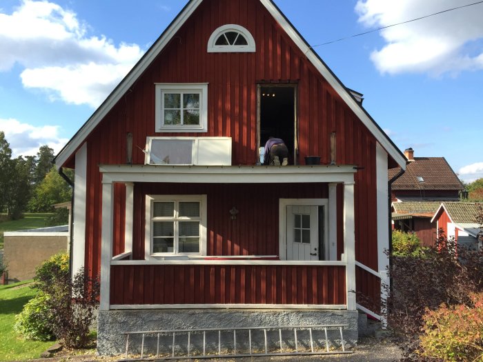 Person bygger balkonggolv på rödmålat hus, befintligt balkongräcke och plåttak synligt.