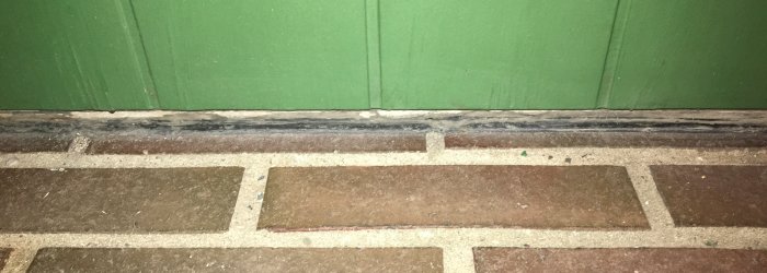 Slitet tröskelområde vid en ytterdörr med gammal svart fogmassa och synlig träskada.