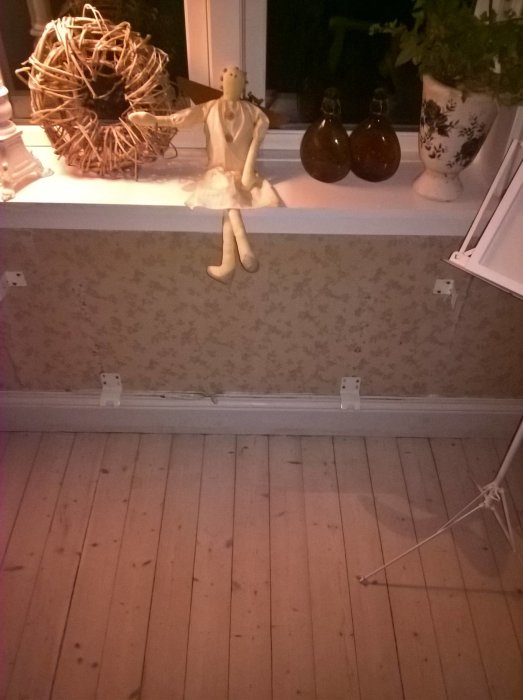 Vägg med borttagna oljeelement, synliga fästen och elledning på trägolv, dekor med docka och krukväxt.