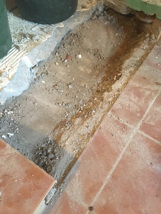 Opbrudt kældergulv med udsatte jord- og stenlag samt synlige keramikfliser; klargjort til udjævning med beton.