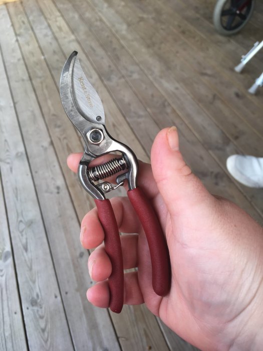 Närbild på en ny sekatör i en hand, mot träveranda bakgrund, mindre och lättare modell.