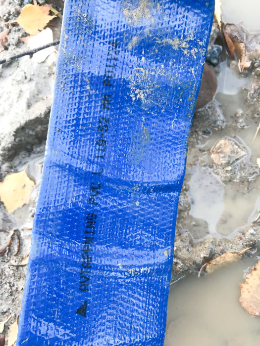 Blå avtappningsslang i lerigt vatten med synliga måttmarkeringar.