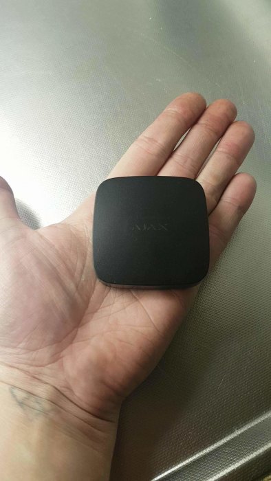 En liten, svart fuktsensor hålls i en hand mot en metallbakgrund.
