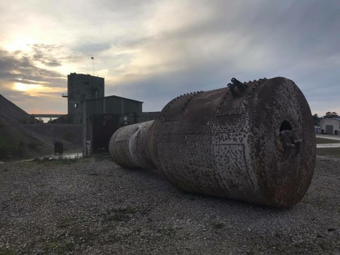 Stor rostig metallbehållare på marken med industribyggnad i bakgrunden vid skymning.